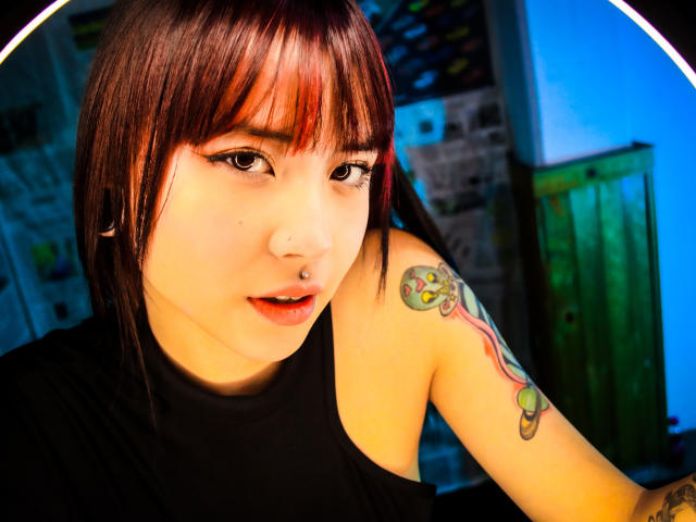 EmmahGreys with red-brown bob, black sleeveless top, nose ring, tattoo on right arm, and black eyeliner, looking at camera with slightly open mouth.