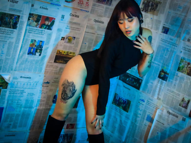 EmmahGreys with dark hair, black long-sleeve top, and black thong, showing a tattoo on her thigh, wearing black knee socks, leaning forward against a newspaper wall.