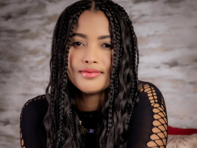 KristalJhonson has long black braids, a cute smile, and wears a black fishnet top, showing her tits subtly through the net. She has light brown skin and pink lips.