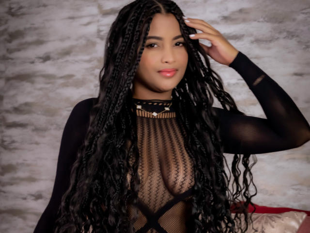 KristalJhonson, con largas trenzas negras, lleva puesto un top de malla negra que deja ver sus pechos, tiene una mano en el pelo y una sonrisa suave, sobre un fondo gris texturizado..
