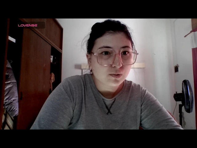 Imsohornyd has pale skin, dark hair in a messy bun, and wears clear glasses with a gray tee. Necklace with a black "X" hangs from her neck.