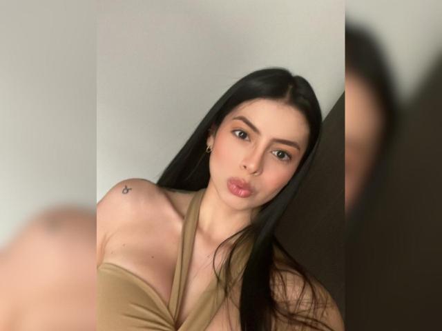 IsabellaJames with long black hair, pouty lips, and a small tattoo on her left shoulder, wearing a tan halter top, showing her boobs in a blurred selfie.