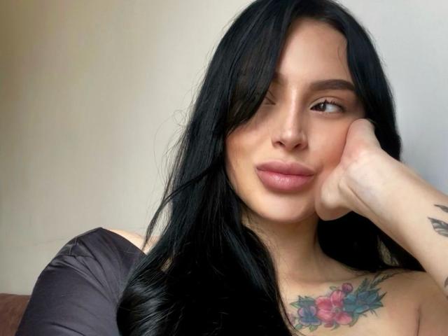AprilTheOne, avec ses longs cheveux noirs, cligne de l'œil, a sa main sur sa joue, et montre un tatouage de fleurs colorées sur sa poitrine. Elle porte un haut noir à une épaule..