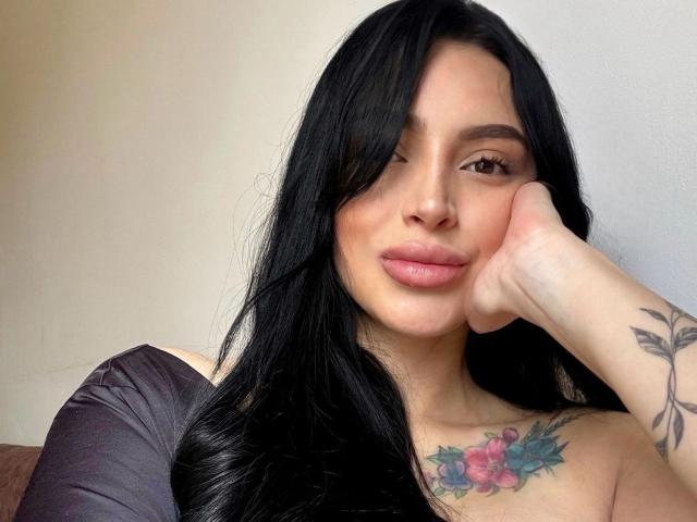 AprilTheOne, com cabelo preto, piscando, com a cabeça apoiada na mão, mostrando uma tatuagem de flores rosa e azul no peito, vestindo uma blusa preta, parecendo adorável..