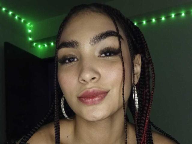 LylaPeach has medium brown skin, thick black eyebrows, long red-braided hair, and wears silver hoop earrings with green string lights in the background. She's smiling with pink lips.