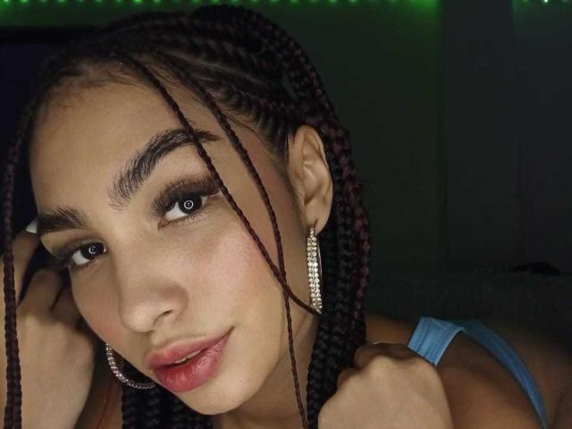 LylaPeach has dark brown braids, thick eyebrows, and wears diamond earrings. She's looking at the camera with slightly parted pink lips, wearing a blue top.