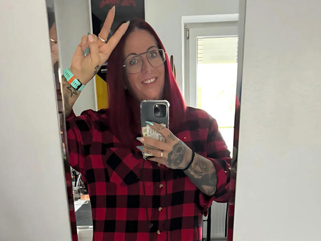 Miss-Ruby-Rubens-hot with red hair, glasses, and tattoos, wearing a red-black plaid shirt, making a peace sign, holding a phone, smiling, showing off her nails.