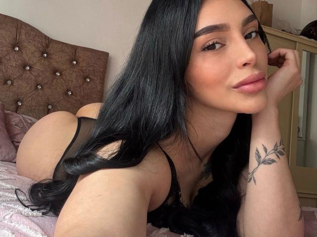 AprilTheOne ha lunghi capelli neri, è sdraiata su un letto con un intimo nero, mostrando il sedere e un piccolo tatuaggio sul braccio destro. Sta guardando la telecamera con un sorriso..