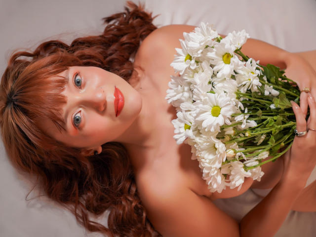 EmilyKlein with red hair, holding white daisies to her bare boobs, looking up with blue eyes, red lipstick, and a ring on her right hand.
