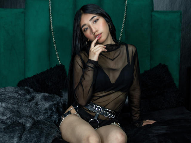 AliGreat with black hair, sheer top, black bra, and belt, finger in mouth, sitting on black fur, showing her tits and pussy, green velvet background, sexy, seductive.