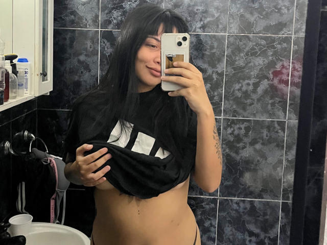 DarkkDesirexx with long black hair, lifting a black shirt to expose her small tits and bare pussy, taking a mirror selfie with a phone, in a dark-tiled bathroom.