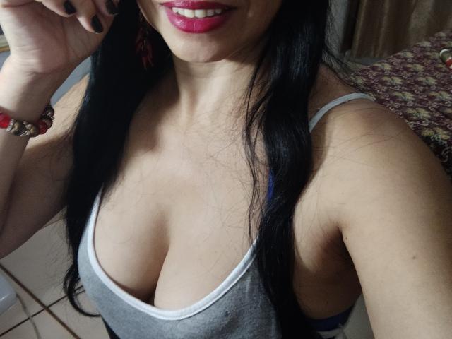 MorenaSexual with long black hair, pink lipstick, and gray tank top showing her big tits and tan skin, wearing red earrings and a bracelet, smiling at the camera.