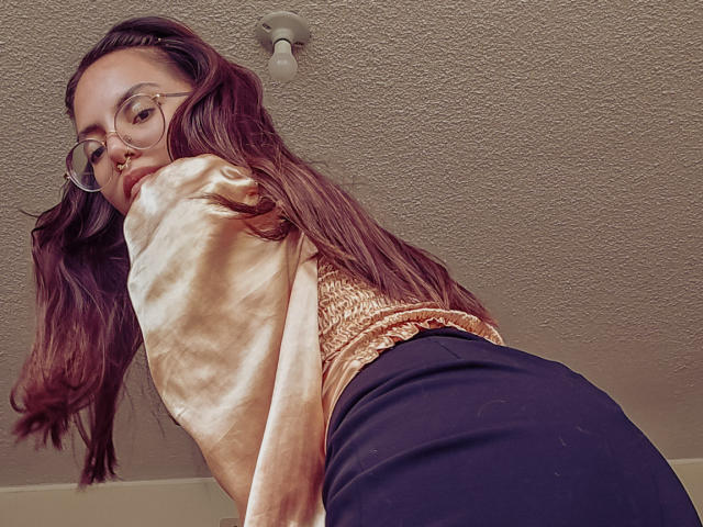 NaturalMariaHun with long brown hair, glasses, tan top, and black pants, bending over, showing her ass, under a textured ceiling with a light fixture.