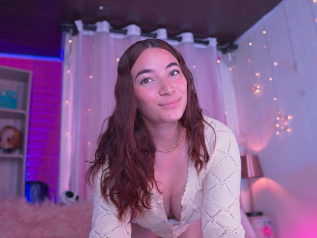 IssaMonterrey with long brown hair, white knit top, and gold necklace, smiling at the camera, showing her cleavage, in a pink-lit bedroom with white curtains and string lights.