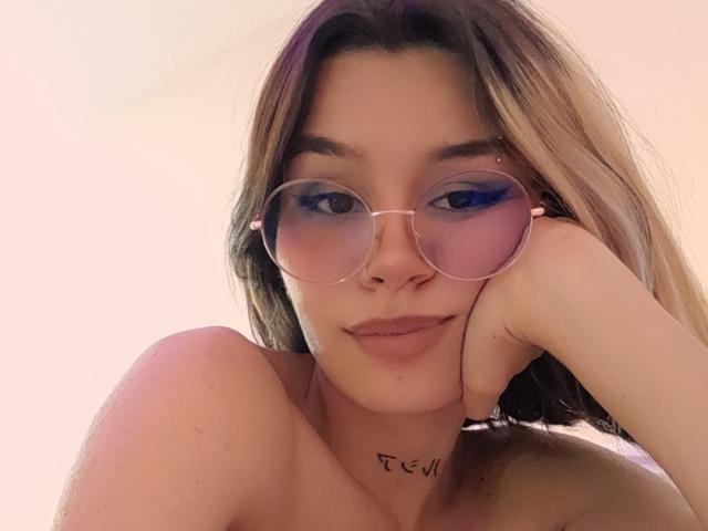 Lena, mit schulterlangem blondem Haar, trägt rosafarbene Sonnenbrillen, blauen Lidschatten und eine Kette um den Hals. Sie legt ihren Kopf in ihre Hand und zeigt ein Tattoo auf ihrer Brust..