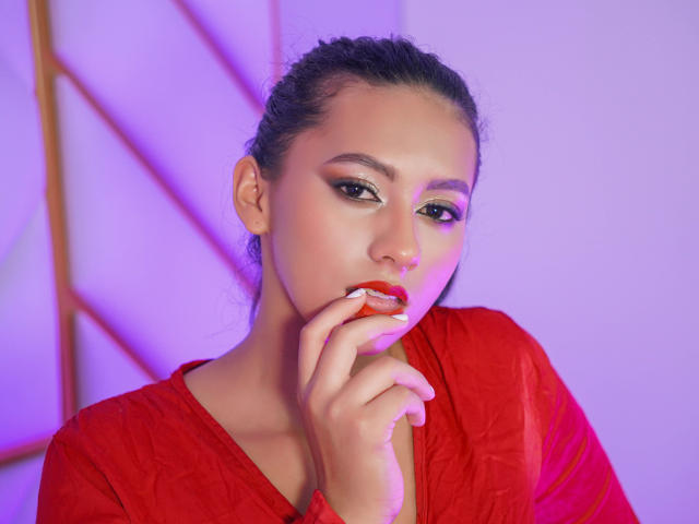 AshleyRosel with red lipstick, green eyeshadow, and red shirt, touching her lips with her finger, looking seductive, purple and pink lighting, dark hair in a ponytail.