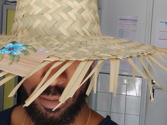 Kelkeu trägt einen breitkrempigen Strohhut mit Fransen und einem blauen Blumenaufkleber, der ihr Gesicht bedeckt. Sie hat einen Bart und trägt ein dunkelblaues Hemd..