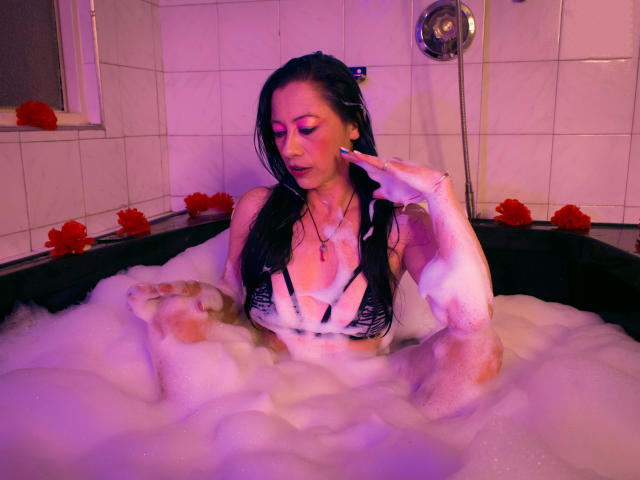 Noemi : La plus belle avec ses cheveux noirs mouillés, portant un soutien-gorge en dentelle noire, assise dans un bain moussant, ses seins couverts de mousse, une main touchant sa joue, des pétales de rose rouge autour d'elle..
