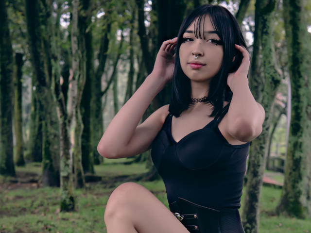 MillaHottLove69 has black hair, a nose ring, and a choker, wearing a black tank top, hands in her hair, forest background, visible tits, cute, sexy, and confident.