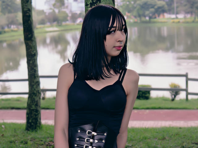MillaHottLove69 mit schwarzem Bob, Nasenring, schwarzem Tanktop und schwarzem Korsett mit Schnallen, steht neben einem Baum und zeigt kleine Brüste und ein süßes Gesicht..