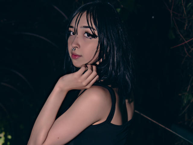 MillaHottLove69 with black hair, nose ring, and eyeliner, looking over shoulder, black tank top, hand on neck, dark background, slightly wet hair, subtle smile, dark lighting.