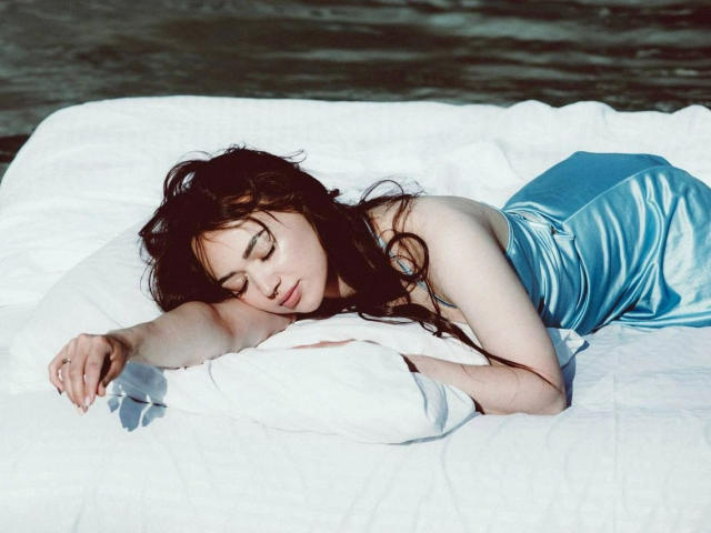 Kristianna, com cabelos escuros e ondulados, olhos fechados, deitada em uma cama branca, vestindo um vestido de cetim azul brilhante, braços sobre um travesseiro branco, aparentando estar relaxada. Água ao fundo..