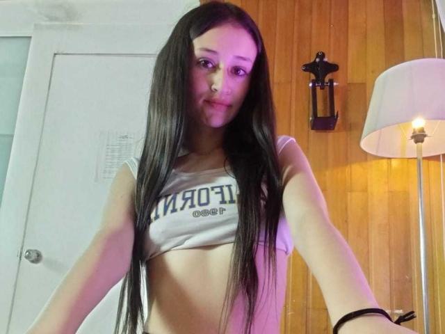 EmilySwiftt met lang zwart haar, lachend, haar tong uitstakend, gekleed in een witte crop top die haar kleine borsten laat zien, en een zwarte armband om haar rechterpols..