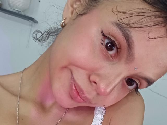 DanesaCronin's face, showing pink-red skin on her neck, brown eyes with long lashes, small silver nose stud, wet curly brown hair, and a white lacey bra strap.