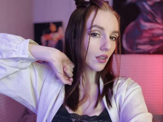 KatKitty with brown hair, white shirt, and black lace top, has her hand in her hair, showing her cute face and slightly open mouth. Soft pink and purple lighting.