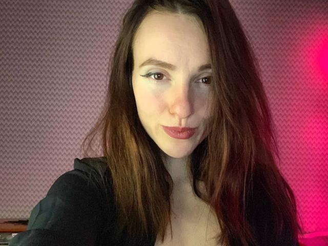 KatKitty with long brown hair, pale skin, and subtle eyeliner, giving a pouty expression, wearing a black top, red light on the right. Boobs partially visible.