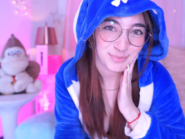 IssaMonterrey with glasses, blue and white furry hood, hand on cheek, smiling, pink and purple lighting, brown hair, red bracelet, stuffed toy in background.