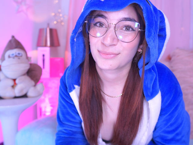 IssaMonterrey with brown hair, glasses, and a blue and white animal hoodie, smiling, pink and purple lighting, small gold necklace, and a stuffed monkey on a white table.