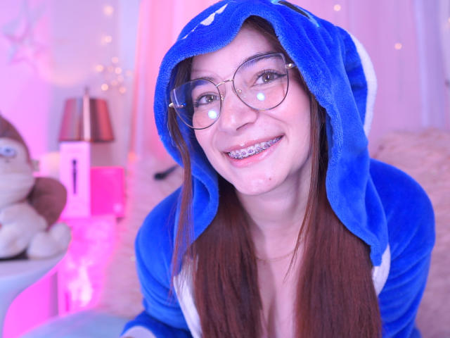 IssaMonterrey in a blue plushie hoodie with a cute smile, glasses, and braces, brown hair, pinkish lighting, and a stuffed toy in the background. Her tits are small.