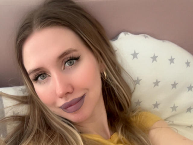 SannyManny has light brown hair, green eyes, and purple lipstick. She's wearing a yellow shirt and gold earrings, lying on a star-patterned pillow, smiling softly.