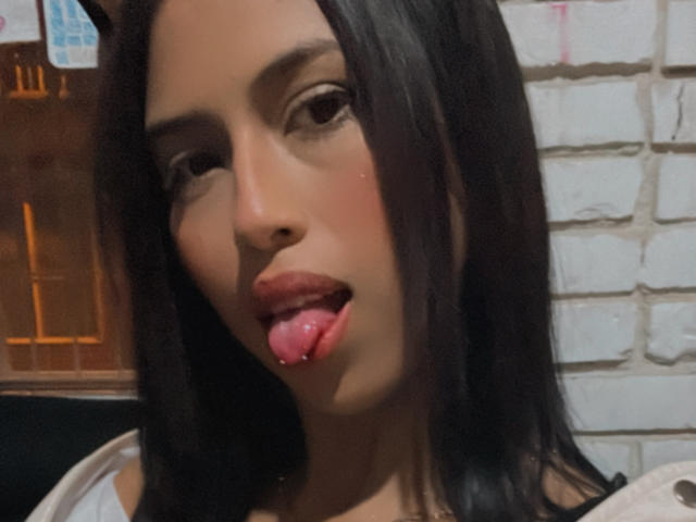 Il viso di MyaaTyler, con i suoi occhi marroni e i capelli neri e lisci, la lingua fuori con un piercing rosa, indossa una maglietta bianca e guarda verso la fotocamera..