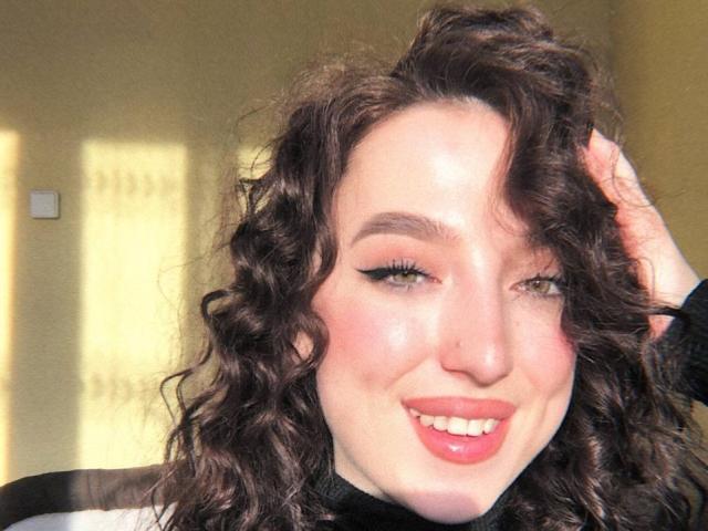 Curly-haired MonAmourR with green eyes, black eyeliner, pink lipstick, and a cute smile, holding her hair, looking directly at the camera, in bright sunlight.