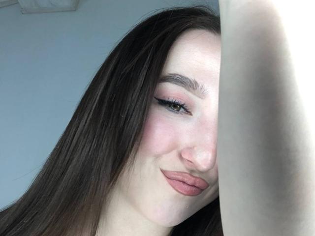 MonAmourR has pale skin, dark brown straight hair, pink eyeshadow, black eyeliner, pink lips, and a slight smile. Her arm is raised, partially blocking the view.
