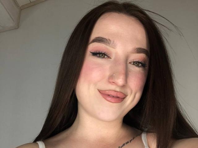 MonAmourR a de longs cheveux bruns, un blush rose et un maquillage subtil avec un eye-liner ailé. Elle sourit, avec un rouge à lèvres rose, et on peut voir un nez mignon et un tatouage sur son côté droit..