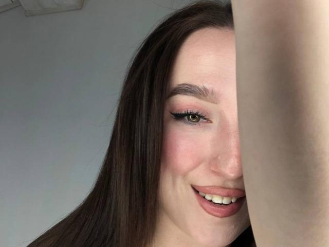 MonAmourR has long brown hair, pink eyeshadow, and is smiling with pink lipstick, showing her white teeth, and has a small nose ring. Her arm is raised to the side.