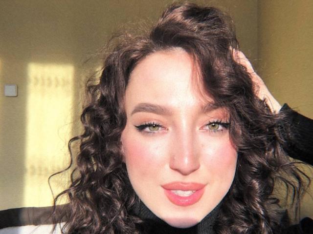 Curly brown-haired MonAmourR with green eyes, pink lips, and black turtleneck, hand in hair, smiling softly, sunlit face, subtle makeup, natural light.