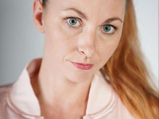 13-Mel-hot with blue eyes, pink nose ring, and pink lips, wearing a pink collared shirt, has long red hair and a slightly smirking expression.
