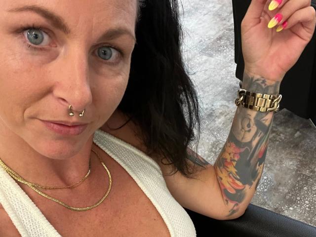 13-Mel-hot with blue eyes, nose ring, and white top, showing tattooed arm with colorful female portrait, and yellow nails, wearing gold chain and watch.