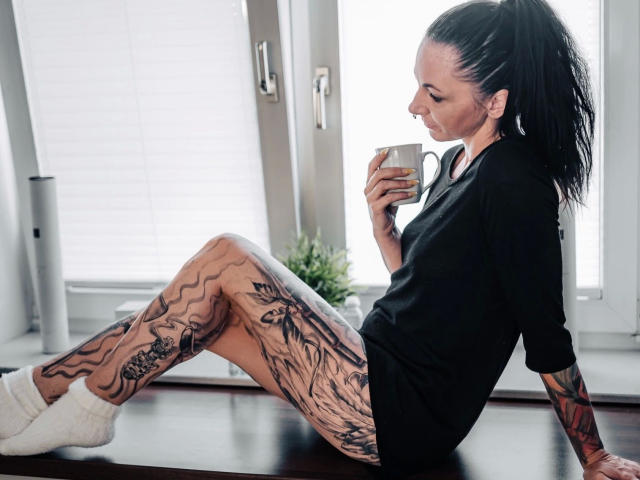 13-Mel-hot with tattoos on legs and arms, black shirt, white socks, sitting on counter, holding white mug, black hair in ponytail, looking down, in daylight room.