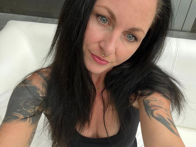 13-Mel-hot has black hair, blue eyes, nose ring, pink lipstick, black tank top showing her boobs, tattoos on both arms, and is smiling at the camera.