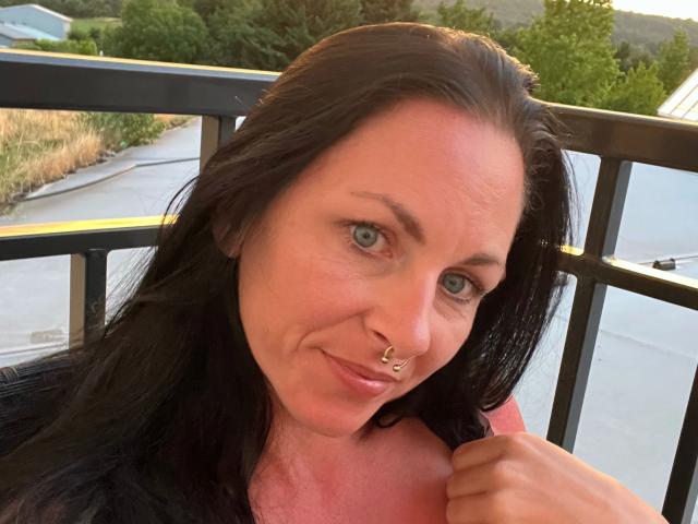 13-Mel-hot with black hair, blue eyes, nose ring, slight smile, pink shirt, holding collar, sunlight, outdoors, black metal railing in background, green trees, calm expression.