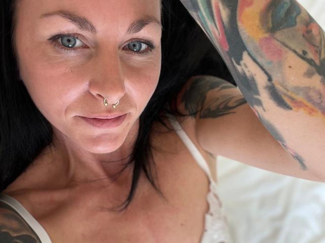 13-Mel-hot with blue eyes, black hair, nose ring, and tattoos on her arm, wearing a white lace top, looking up at the camera.
