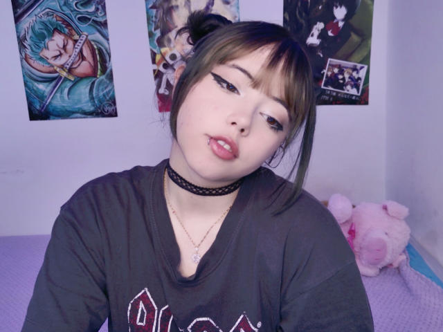 Arieyaara with light skin, brown hair in pigtails, black eyeliner, pink lipstick, black choker, black shirt, pink stuffed pig, anime posters on white wall, purple bed.