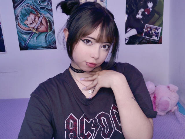 Arieyaara with dark hair in pigtails, wearing a black choker and "Pigros" shirt, has a ring on her finger and a lip piercing, with a pink stuffed toy.