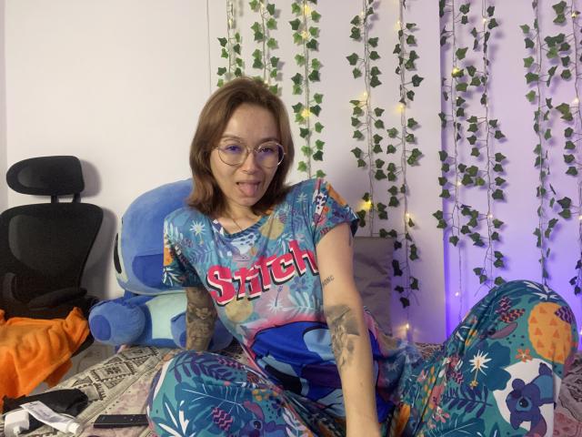 SarahRousen with glasses, brown hair, and tattoos, wearing colorful "Sitch" shirt and pants, sticking out tongue, sitting on bed, surrounded by fairy lights and vines.