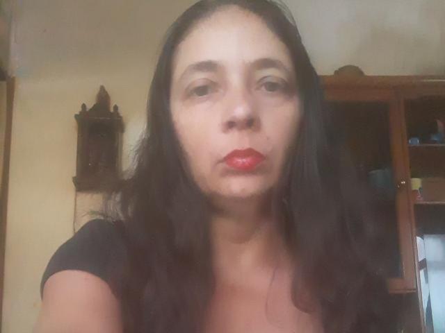 BettyHotBabe with red lipstick, dark wavy hair, and a serious expression, wearing a black top, in a slightly blurred image. Her boobs are average-sized and she's pouting.
